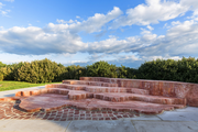 Image of the Bondi Memorial, a red travertine sculpture in the shape of six tiered levels or steps each representing a band in the pride flag. Behind the sculpture is natural coastland shrubs and a view towards Bronte and Clovelly.