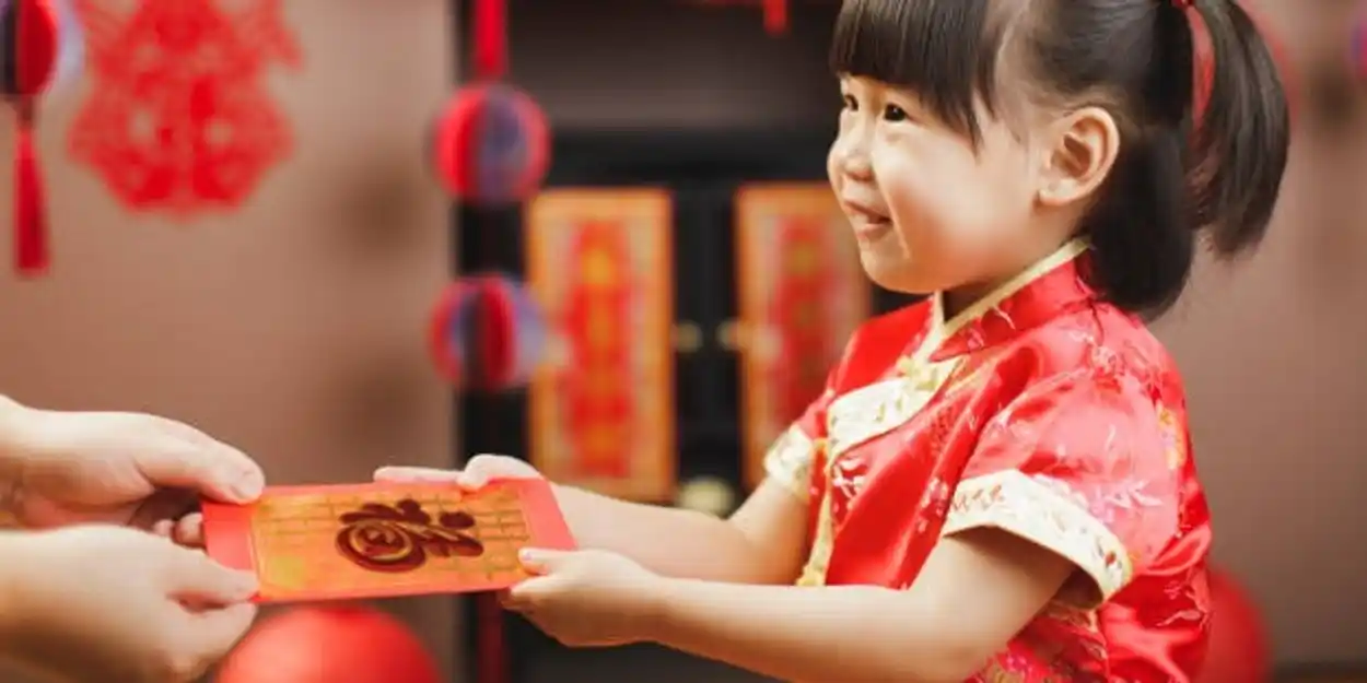 A photograph of a young child dressed in a red and gold gown. An adult is handing her a decorated red envelope.